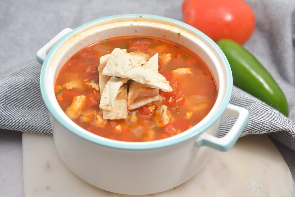 A white bowl of chicken tortilla soup topped with tortilla strips, placed on a cloth with a tomato and a jalapeño in the background.