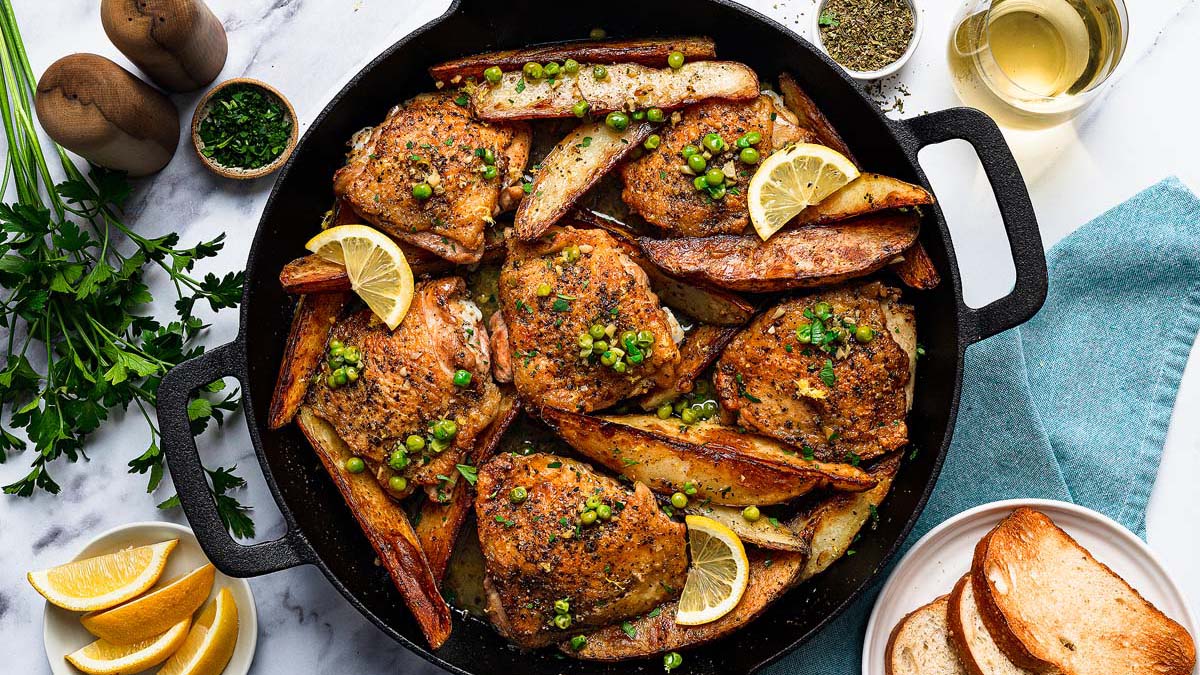 A skillet filled with roasted chicken thighs, potato wedges, green peas, and lemon slices; garnished with herbs, with bread and lemon wedges on the side.
