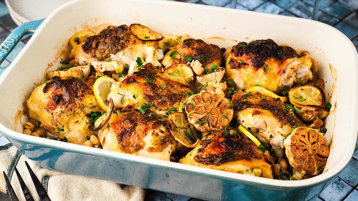 A baking dish filled with roasted chicken thighs, halved heads of garlic, lemon slices, and herbs.