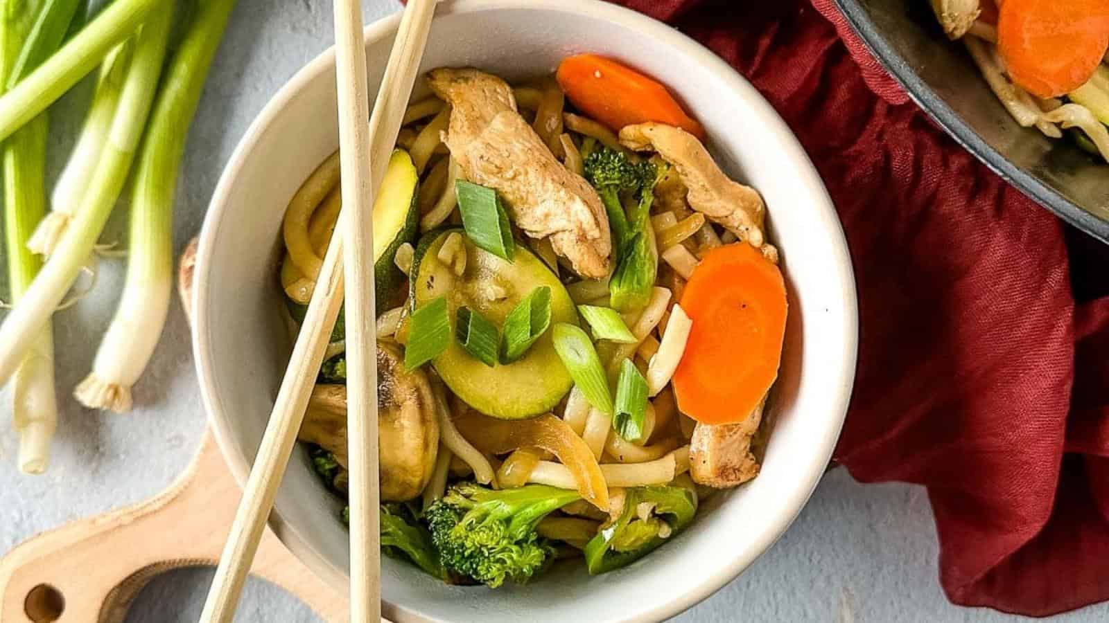 A bowl of stir-fried noodles with chicken, zucchini, broccoli, carrots, and green onions, with chopsticks resting on the bowl.
