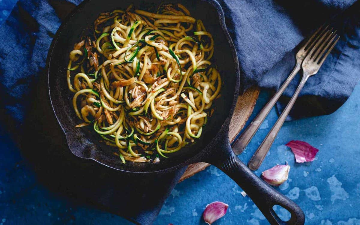 A cast iron skillet filled with zucchini noodles sits on a dark cloth, with two forks and garlic cloves nearby on a blue surface.