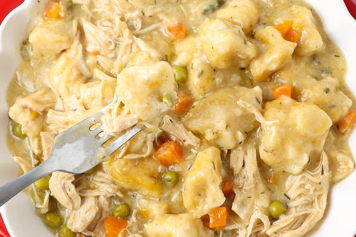 A close-up of chicken and dumplings with shredded chicken, vegetables, peas, and carrots in a creamy sauce, with a fork holding a bite.