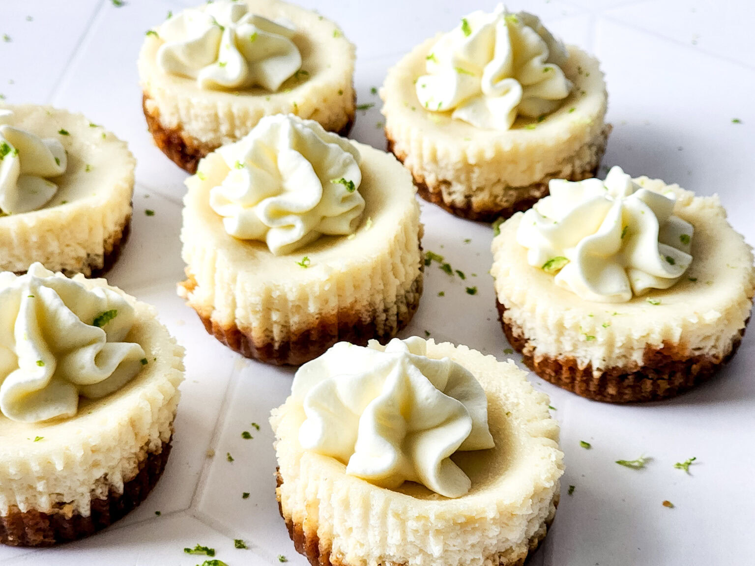 Mini cheesecakes with graham cracker crust, topped with swirls of whipped cream and a sprinkle of lime zest, arranged on a white surface.