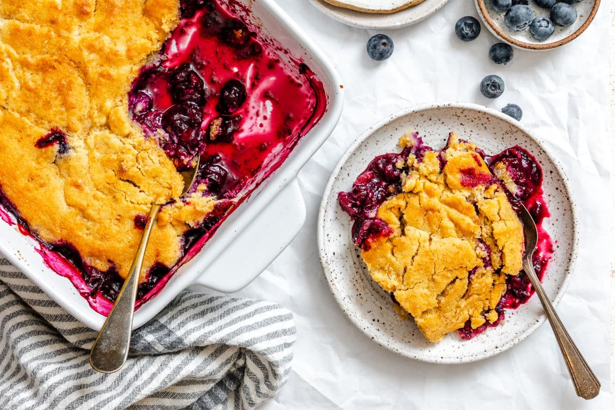 A serving of blueberry cobbler is plated next to a baking dish of cobbler with a serving spoon, surrounded by fresh blueberries on a white surface.