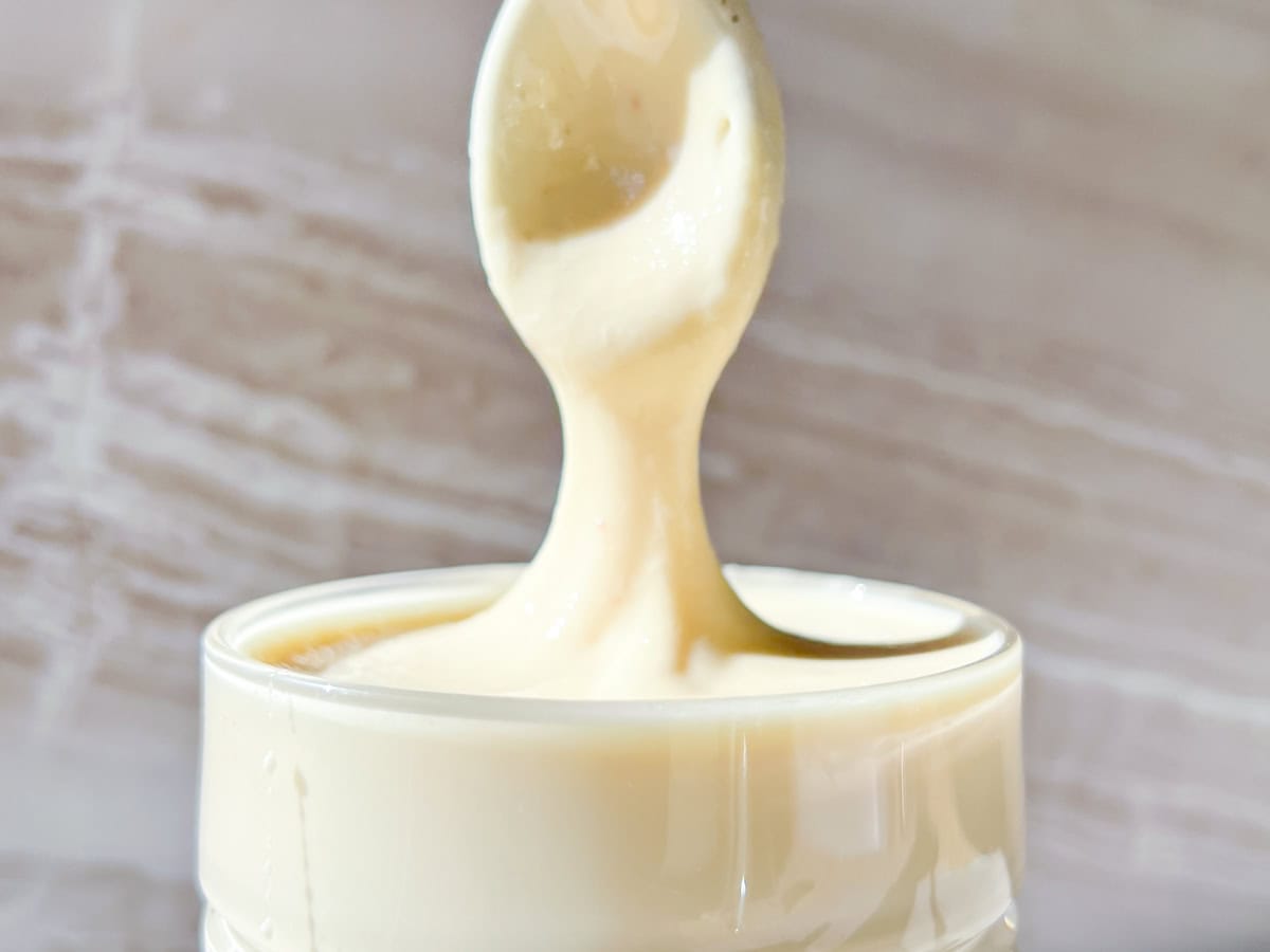 A spoon lifts thick, creamy white substance from a glass jar, with the mixture stretching between the spoon and the jar.