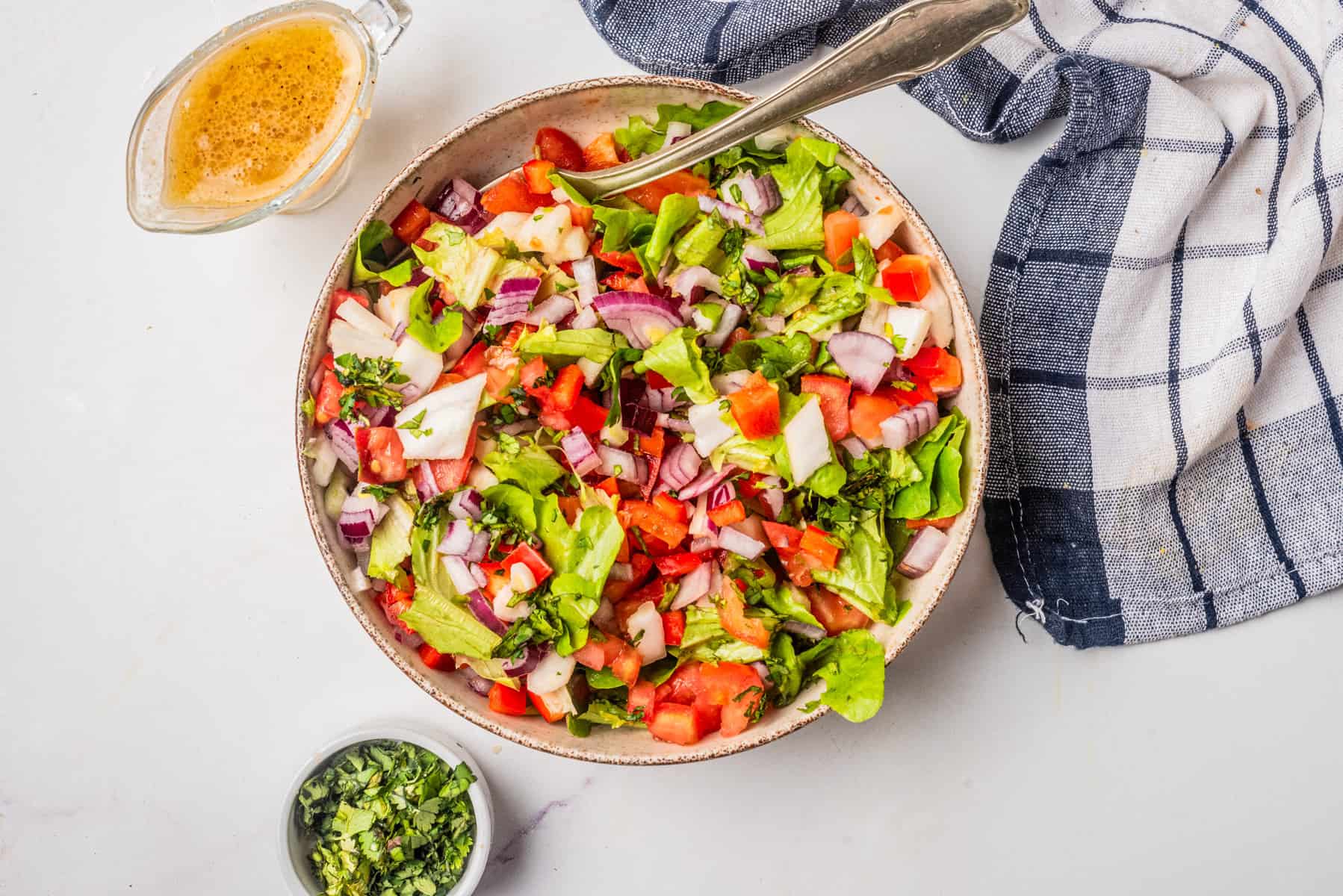 A bowl of chopped salad with lettuce, tomatoes, onions, and herbs next to a small bowl of chopped herbs and a glass container of salad dressing on a white surface.
