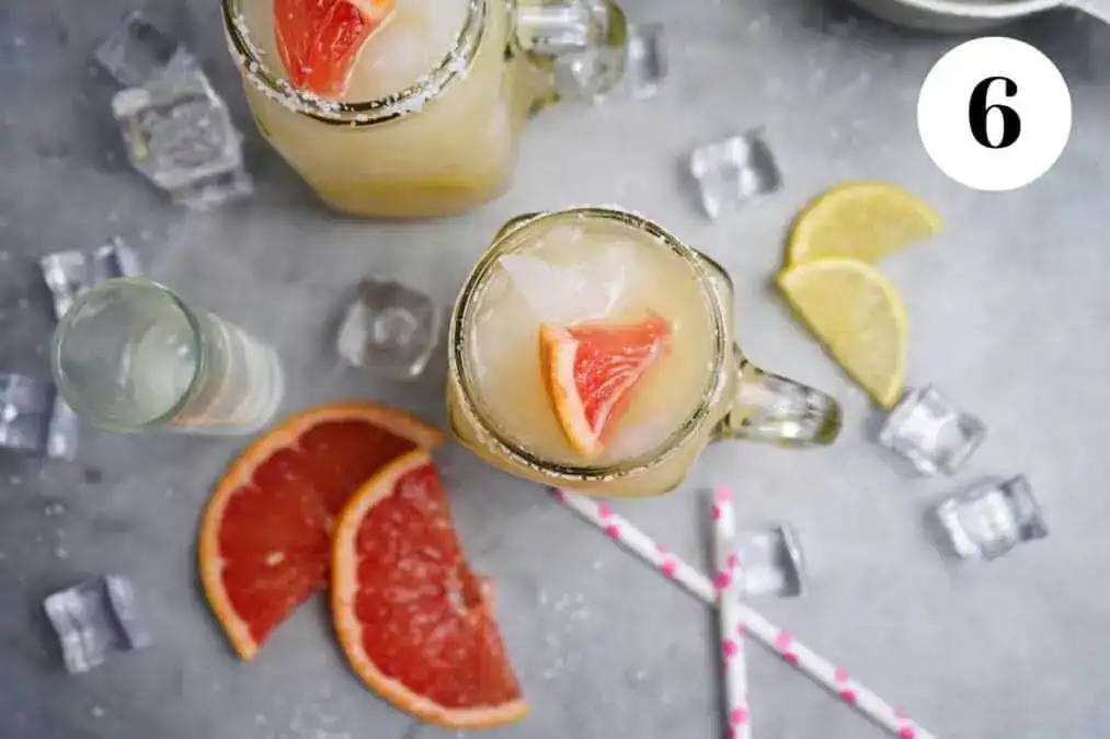 Two mason jar glasses filled with iced grapefruit drinks, garnished with grapefruit wedges, surrounded by ice cubes, lemon slices, and striped straws on a gray surface.