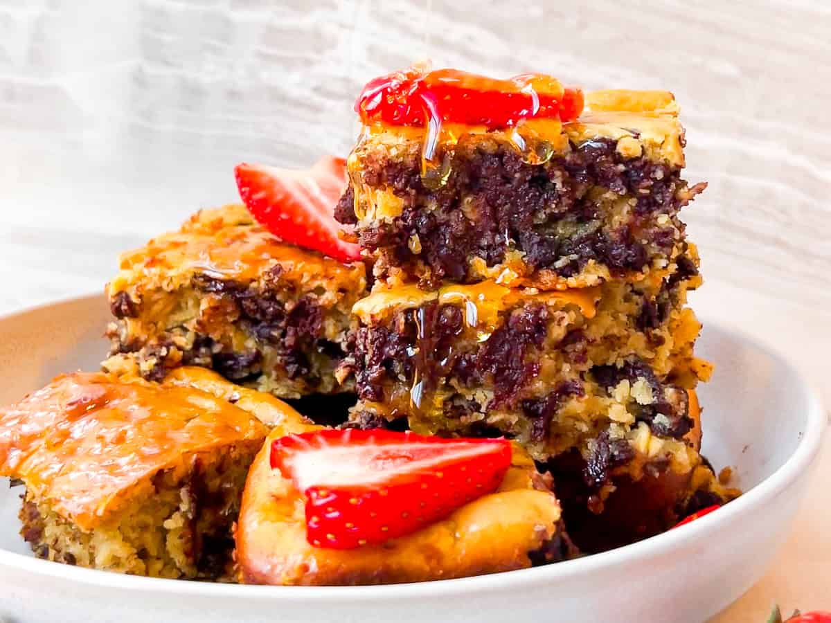 A stack of chocolate chip oatmeal bars topped with sliced strawberries and drizzled with honey, served in a white bowl.