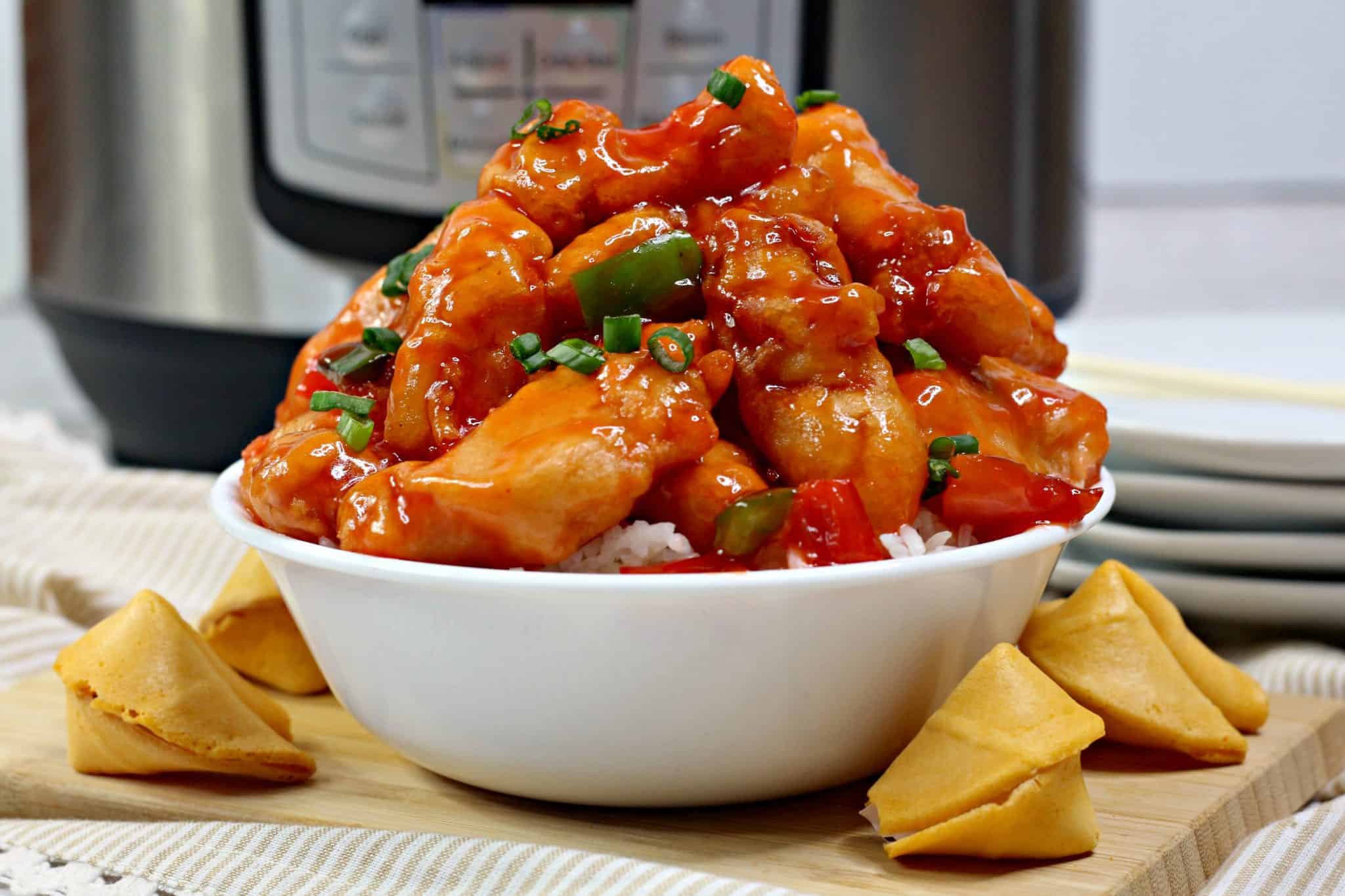 A bowl of white rice topped with sweet and sour chicken, garnished with chopped green onions, surrounded by four fortune cookies on a wooden surface.