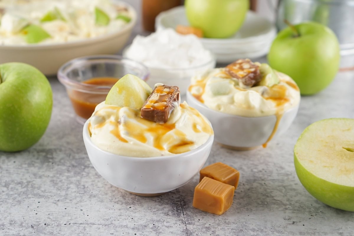 Two bowls of creamy dessert topped with apple slices, caramel sauce, and candy pieces, surrounded by green apples, caramel cubes, and whipped cream.