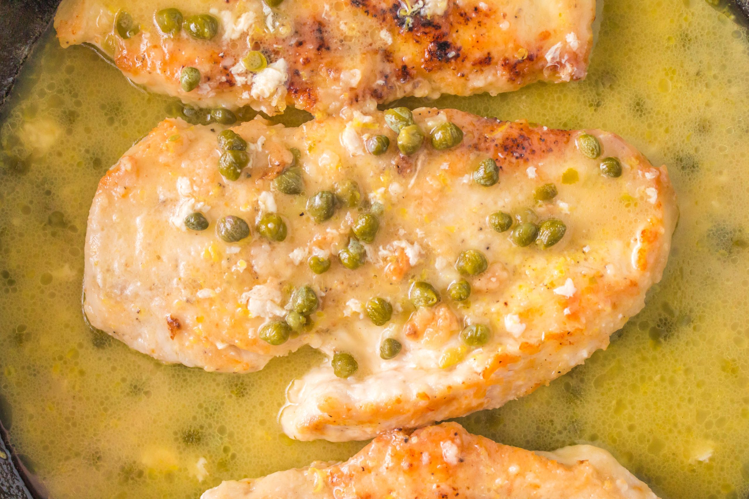 Three pieces of chicken breast in a lemon butter sauce with capers, cooked in a skillet.
