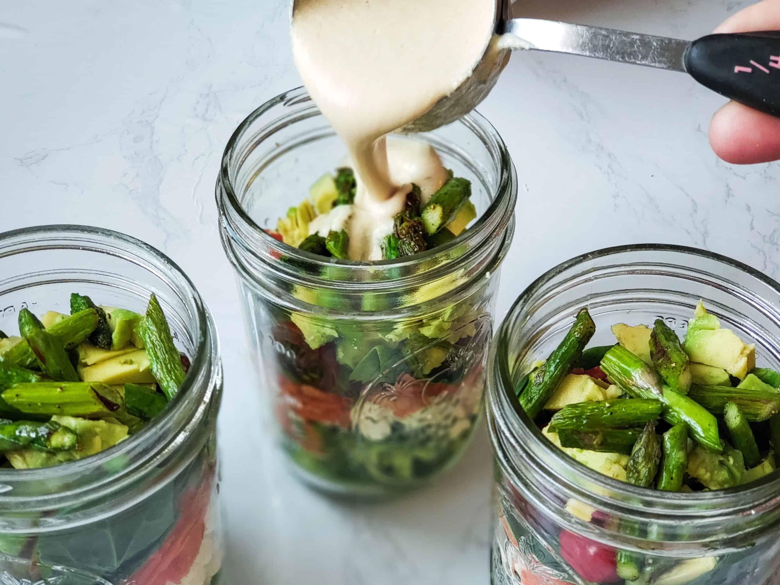 Three glass jars filled with chopped vegetables, with a creamy dressing being poured into the center jar from a metal measuring cup.