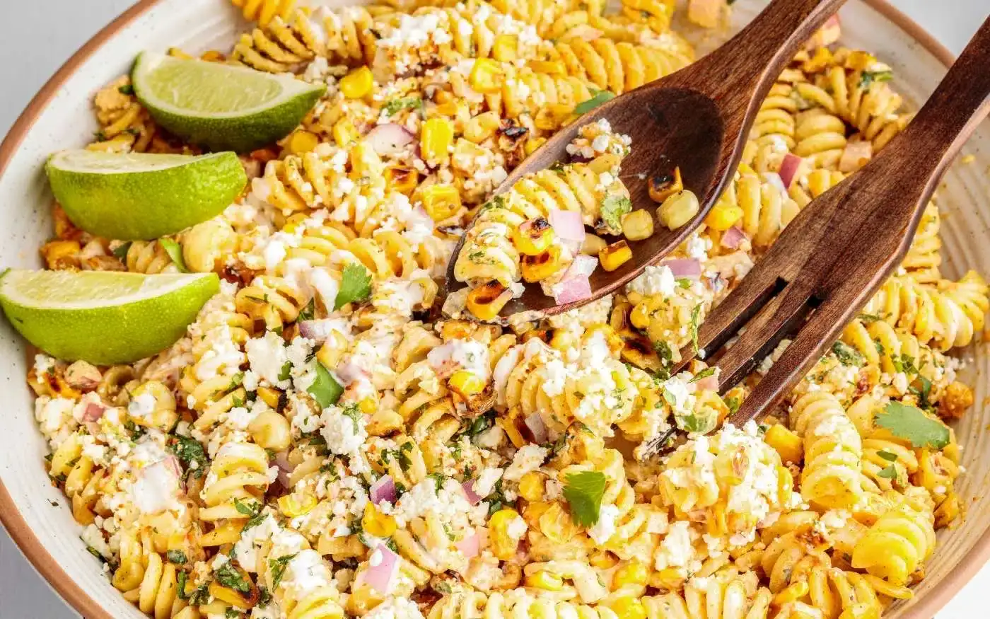 A bowl of rotini pasta salad with corn, red onion, cheese, herbs, and lime wedges, served with wooden utensils.