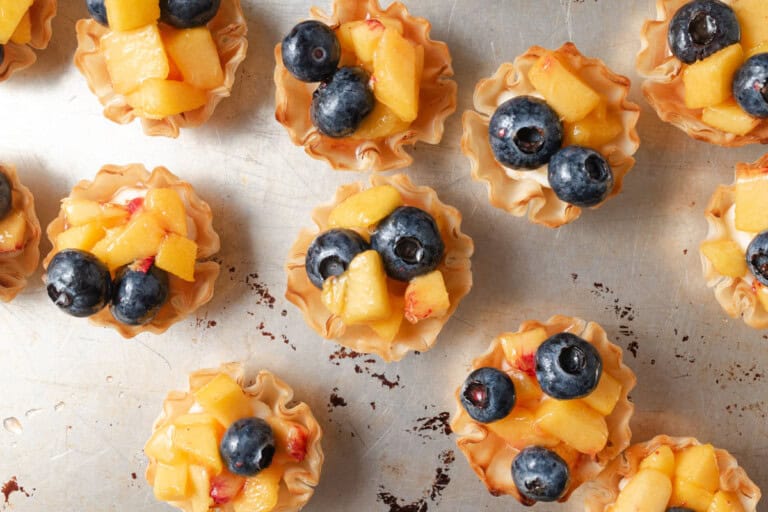 Mini phyllo pastry cups filled with diced peaches and whole blueberries arranged on a metal baking sheet.