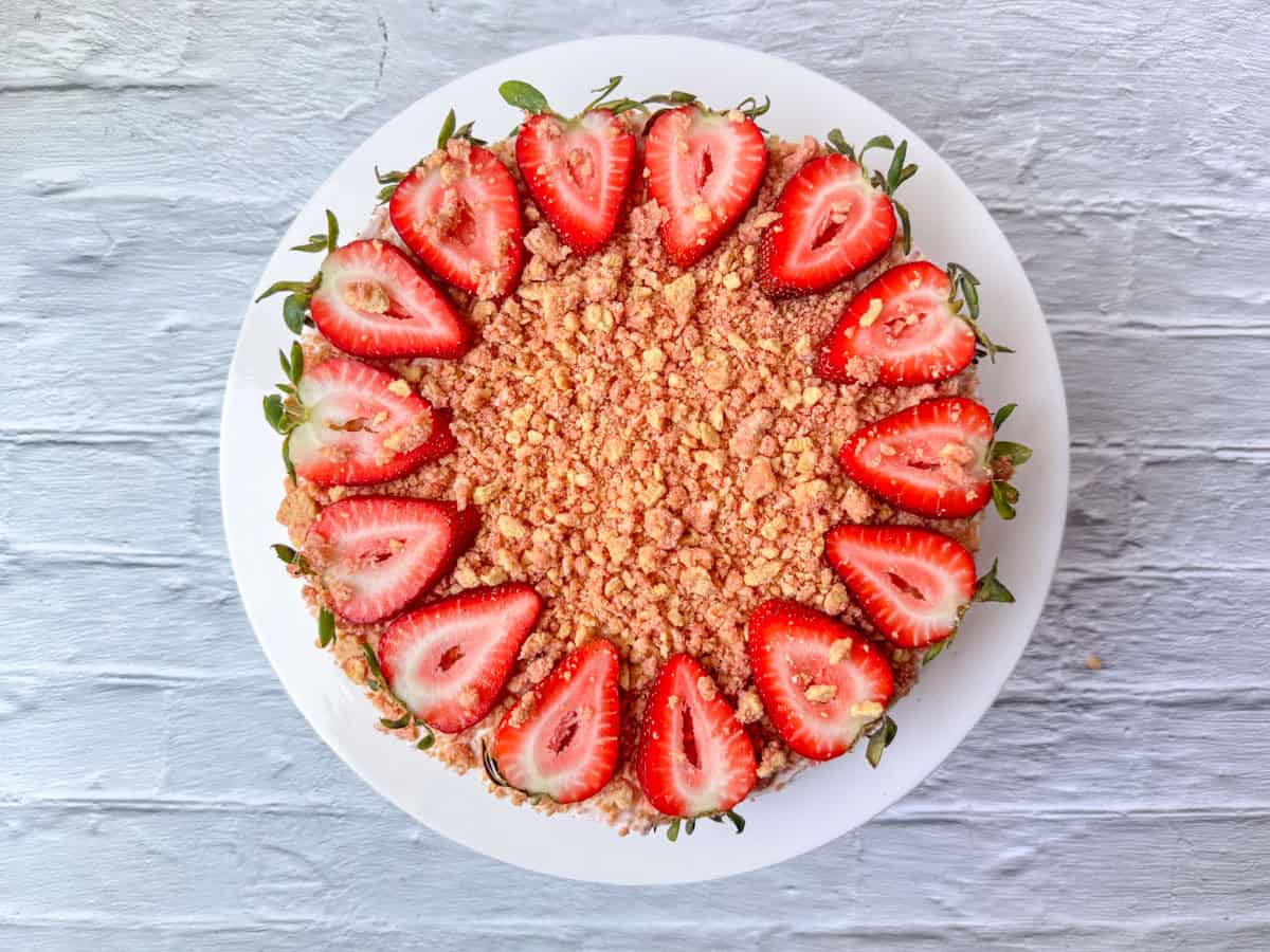 A round cake topped with halved fresh strawberries and a layer of crumbly streusel, presented on a white plate against a light wooden background.