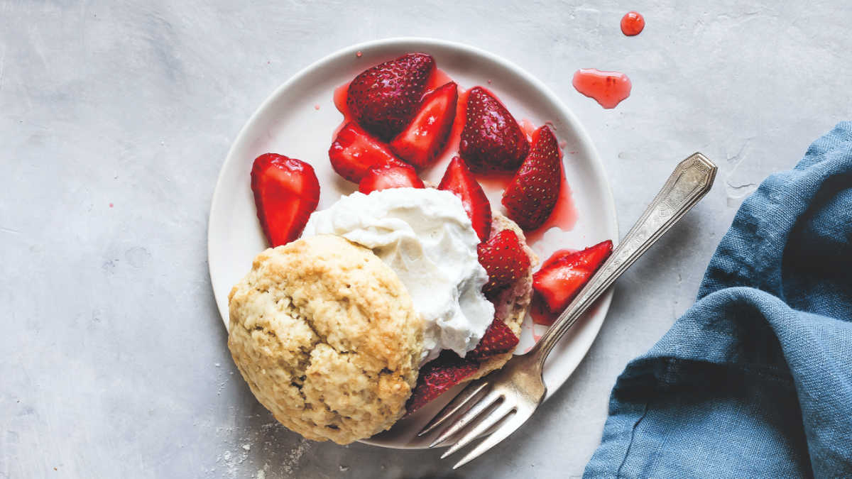 A plate with a biscuit, sliced strawberries, and whipped cream, with a fork beside the food and a blue napkin on the side.