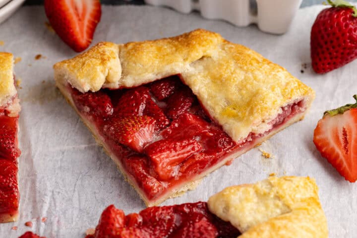 A slice of strawberry galette with a golden-brown crust sits on parchment paper, surrounded by fresh strawberries.