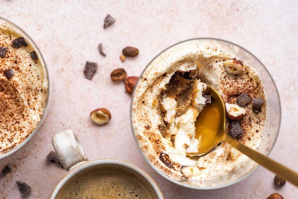 A glass of creamy dessert topped with whipped cream, cocoa, chopped hazelnuts, and chocolate pieces next to a cup of coffee. A spoon is digging into the dessert.