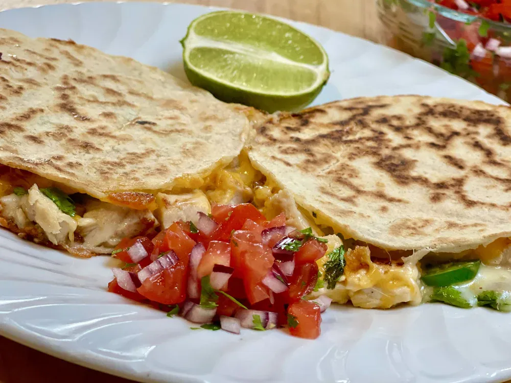 A plate with two grilled quesadillas filled with melted cheese and vegetables, served with a lime wedge and a side of chopped tomato and onion salsa.