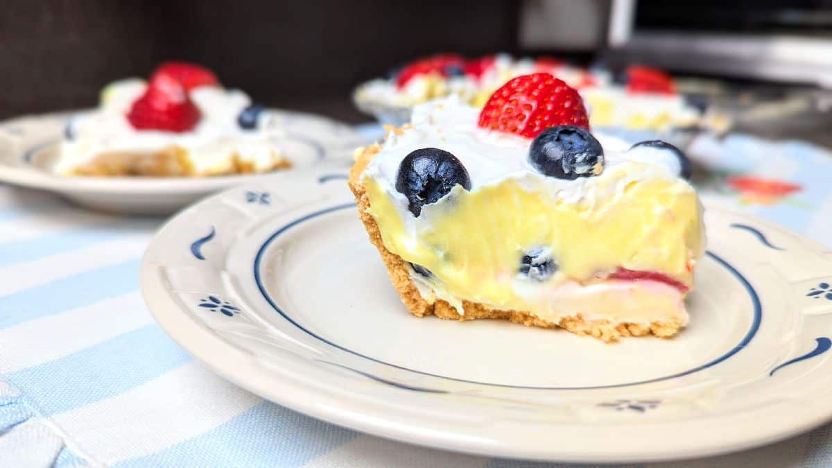 A slice of berry-topped cream pie with graham cracker crust on a plate, featuring blueberries and a strawberry on top.