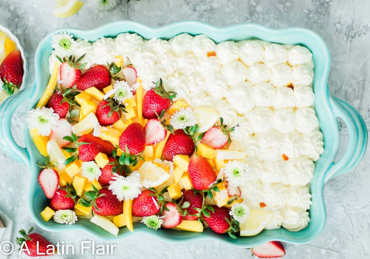 A rectangular dish with half covered in whipped cream dollops and half with strawberries, mango, and citrus slices, garnished with white flowers.