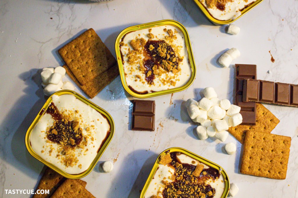 Overhead view of smores-inspired desserts in square dishes, surrounded by graham crackers, chocolate pieces, and mini marshmallows on a white surface.