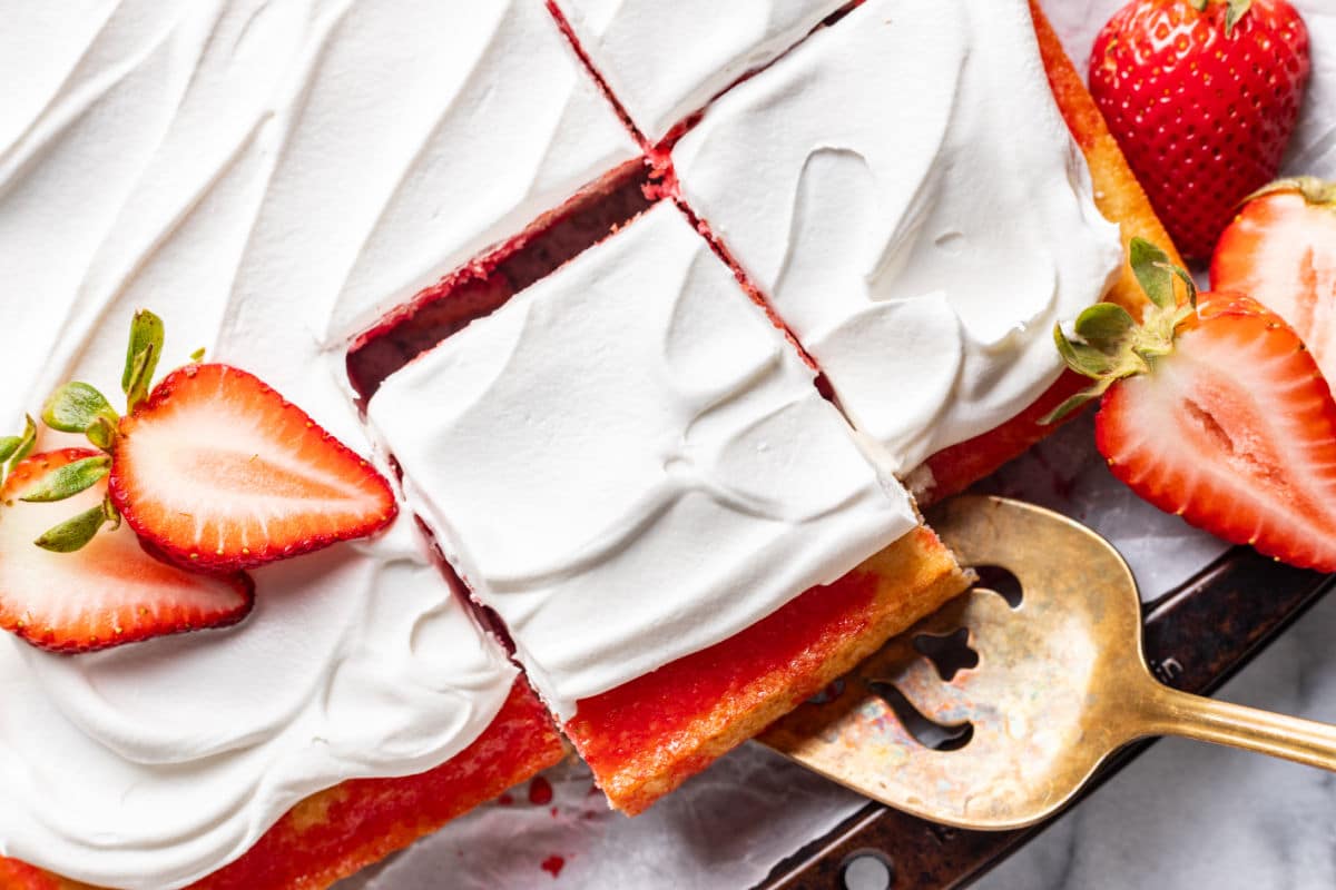 A rectangular strawberry cake with white whipped cream frosting, sliced into squares, and garnished with fresh halved strawberries.
