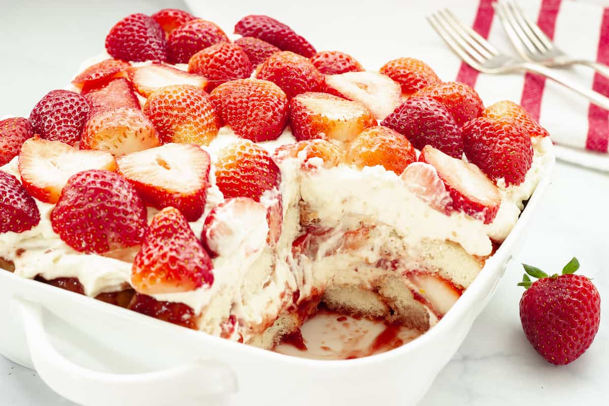 A square dish of strawberry tiramisu with a slice removed, showing layers of cream, ladyfingers, and sliced strawberries. Three forks and a napkin are in the background.