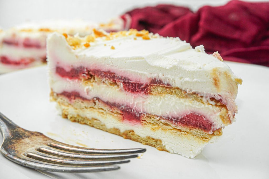 A slice of layered icebox cake with whipped cream, raspberry filling, and graham crackers sits on a white plate next to a fork.