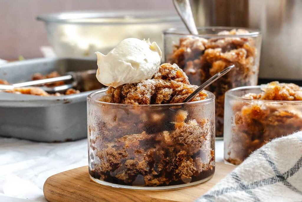 Three glass cups filled with brown apple crisp dessert, one topped with whipped cream, with a baking dish and serving spoon in the background.
