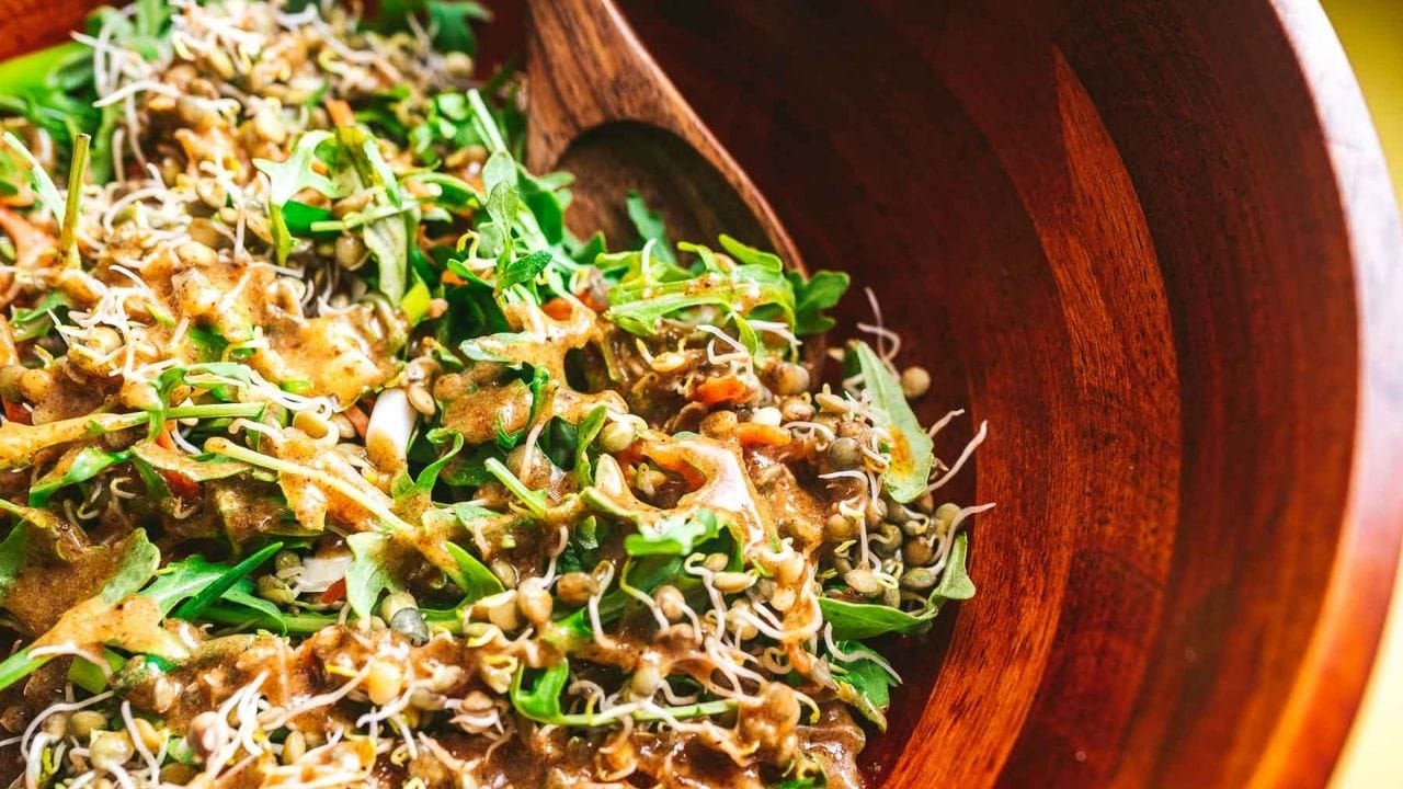A wooden bowl filled with salad greens, sprouts, and a brown vinaigrette dressing, with a wooden serving spoon.
