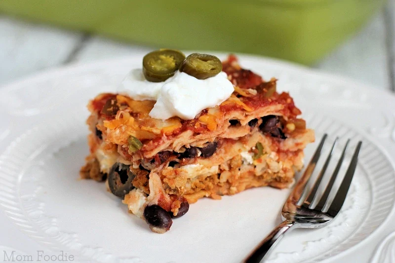 A slice of layered Mexican casserole topped with sour cream and jalapeño slices, served on a white plate with a fork.