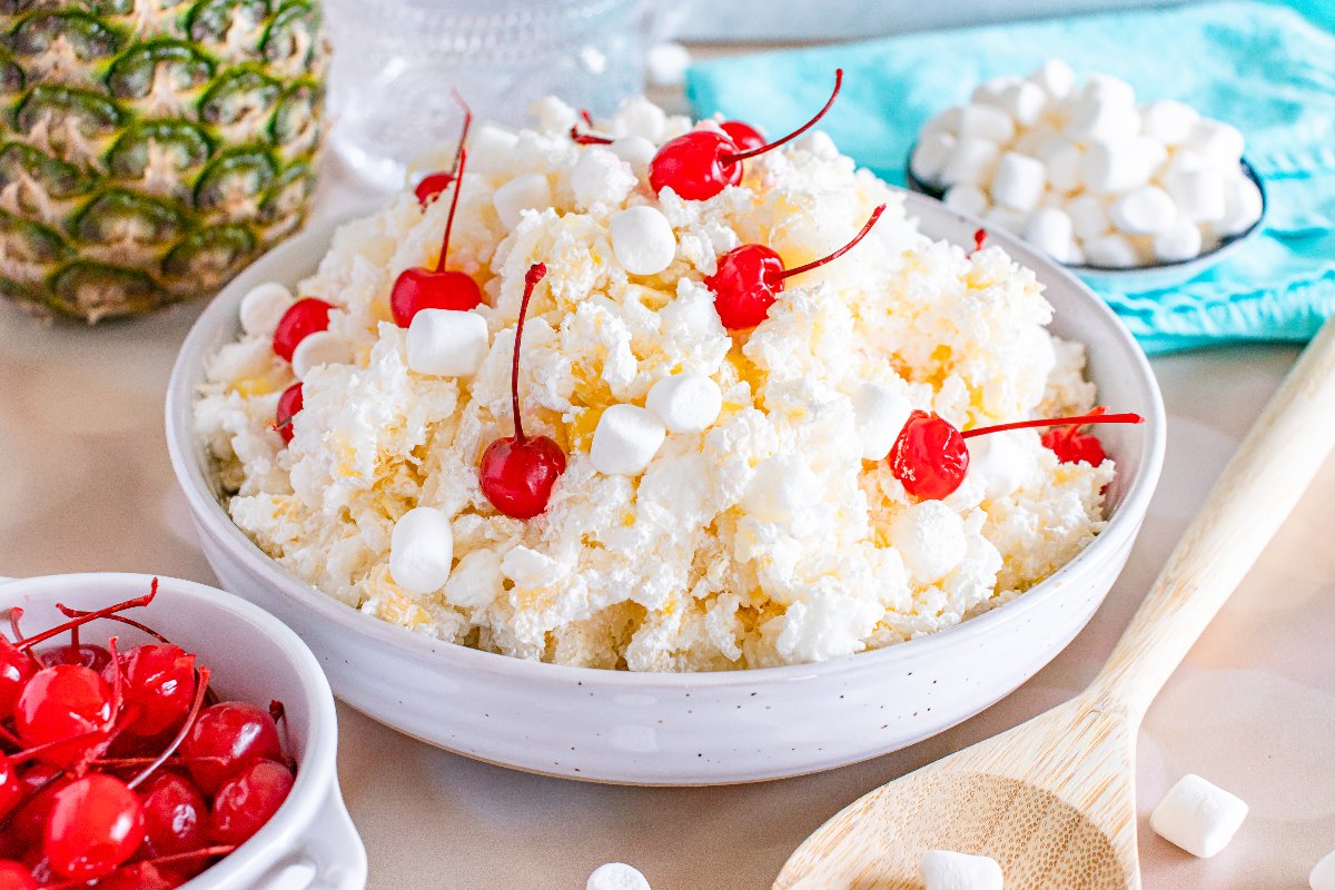 A white bowl filled with a creamy salad topped with maraschino cherries and mini marshmallows, with pineapple and extra cherries in the background.