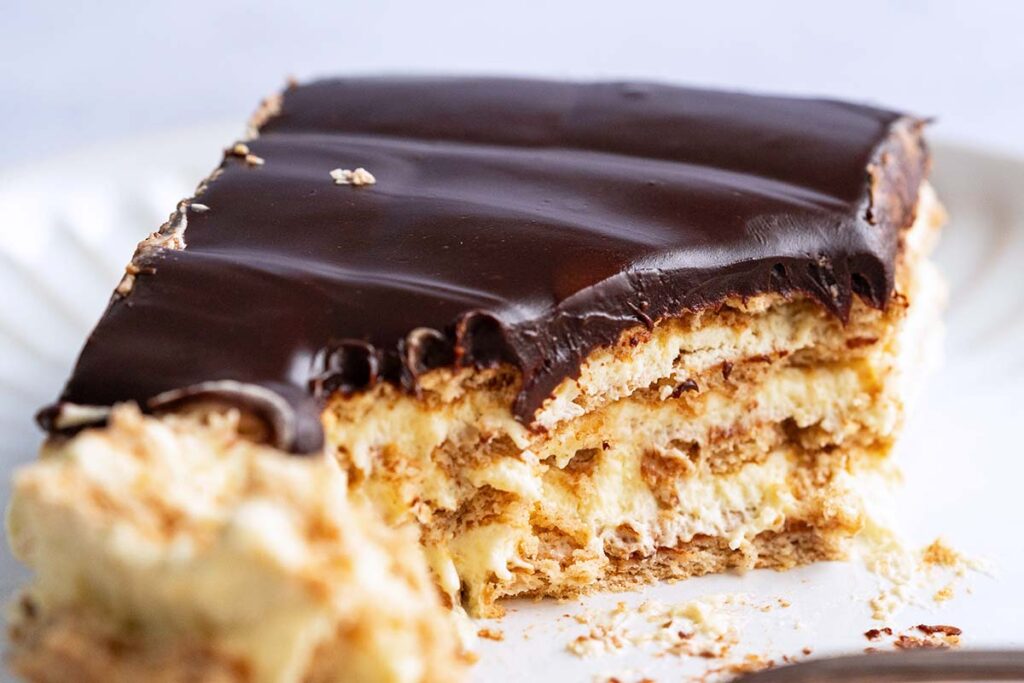 A slice of layered dessert with chocolate topping and creamy filling, partially eaten, on a white plate.