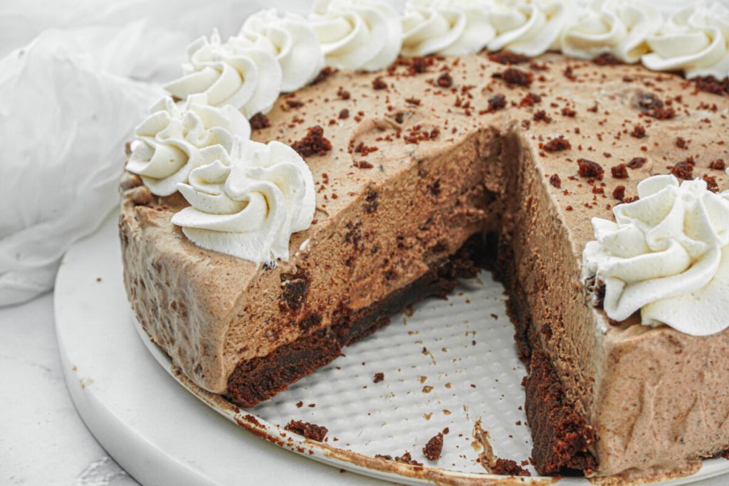 A chocolate mousse cake with a slice removed, showing a layered interior. The cake is topped with whipped cream swirls and chocolate crumbs.