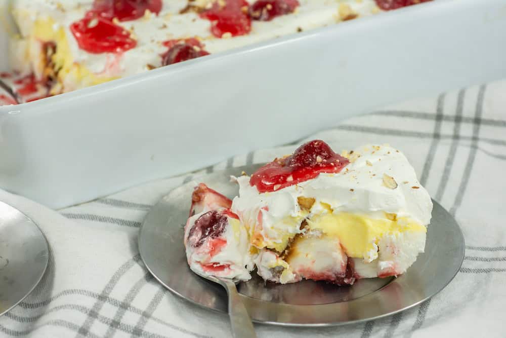 A slice of layered dessert with whipped cream, yellow custard, and cherries on a metal plate, with a baking dish of the same dessert in the background.