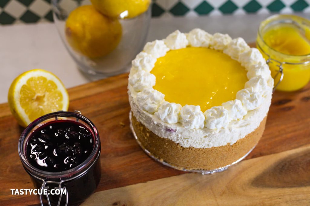 A lemon cheesecake topped with whipped cream and lemon curd sits on a wooden board, next to a jar of berry sauce, a sliced lemon, and a bowl of whole lemons.
