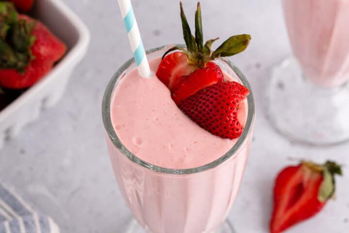 A glass of strawberry milkshake with a blue and white striped straw, garnished with fresh strawberry slices.