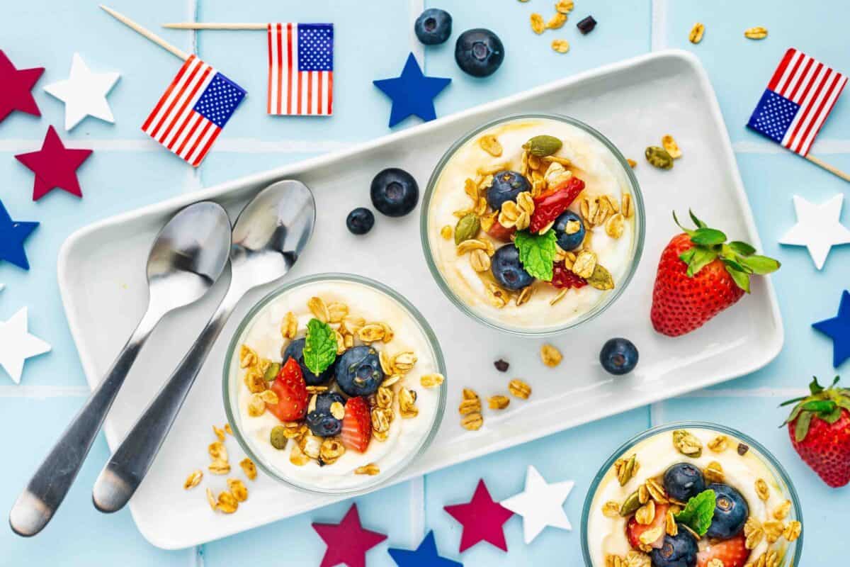 Three bowls of yogurt parfait with granola and berries on a tray, surrounded by American flag toothpicks, red, white, and blue stars, and two spoons on a light blue background.
