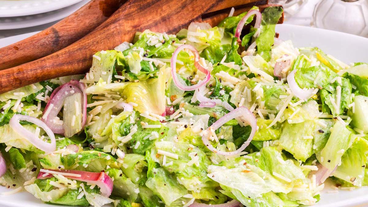 A bowl of green salad with lettuce, sliced red onions, grated cheese, and dressing, served with wooden salad tongs.