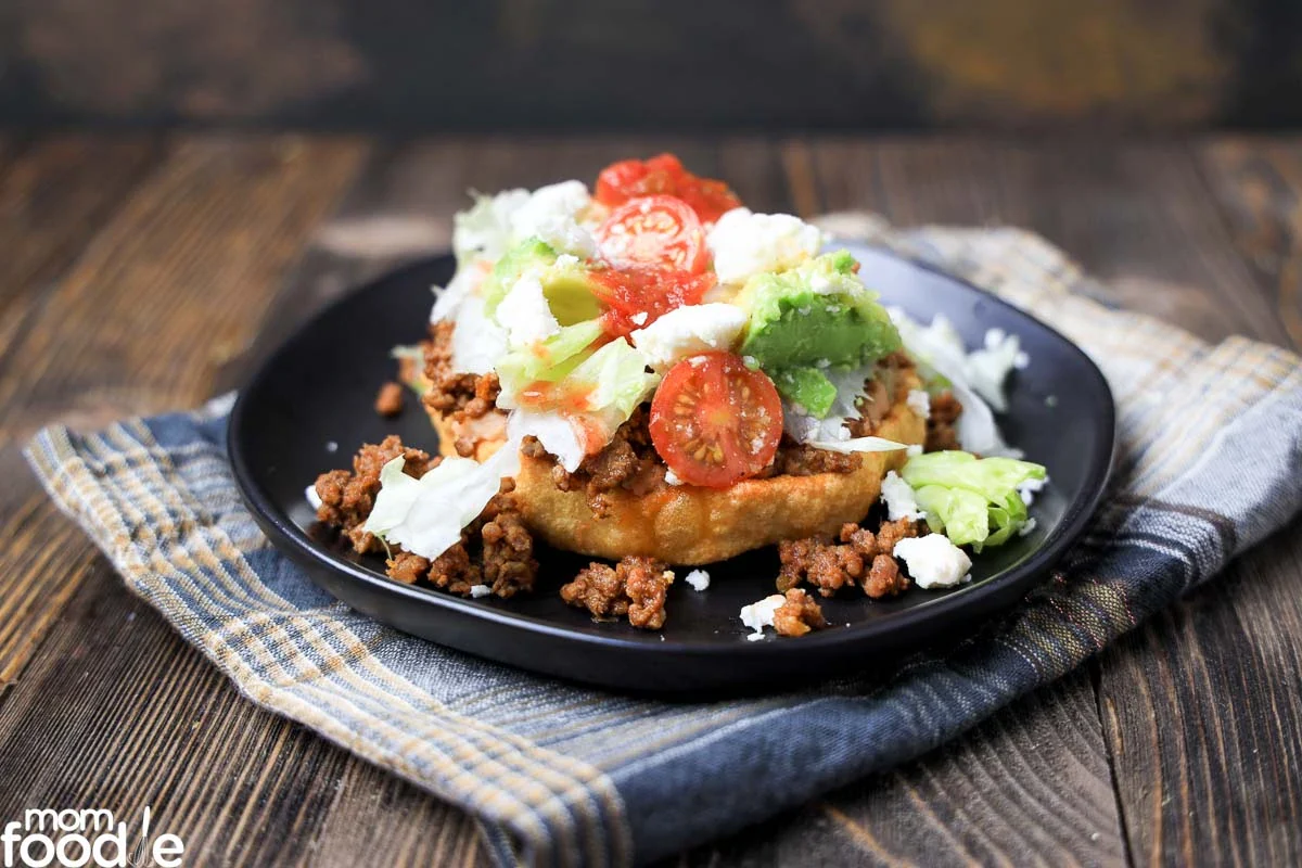 A plate of frybread topped with seasoned ground beef, lettuce, sliced cherry tomatoes, avocado, and cheese, set on a cloth napkin over a wooden surface.