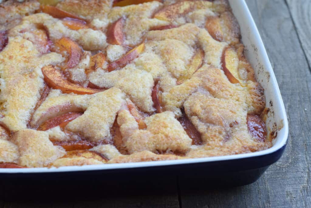 A baked peach cobbler in a rectangular dish sits on a wooden surface, showing a golden, bubbly crust with visible peach slices.