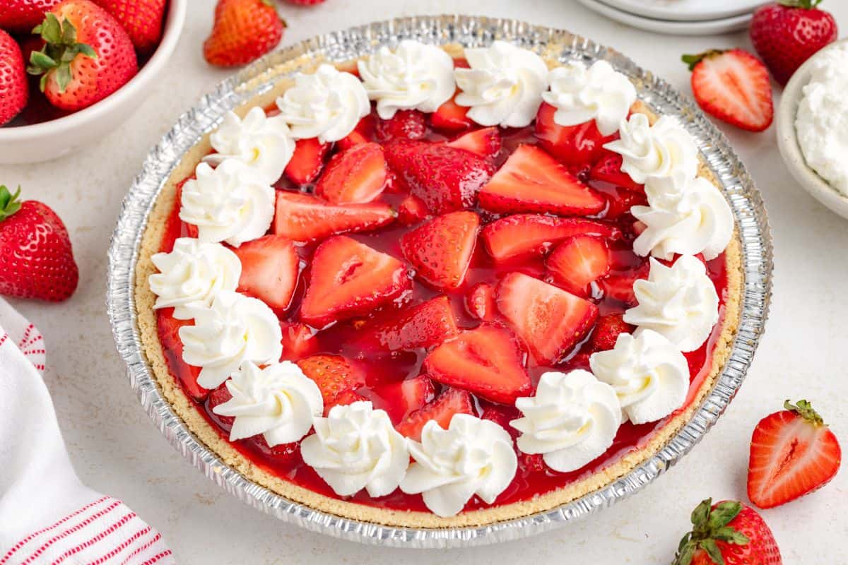 A strawberry pie with a graham cracker crust is topped with fresh strawberries, strawberry glaze, and whipped cream swirls around the edge.