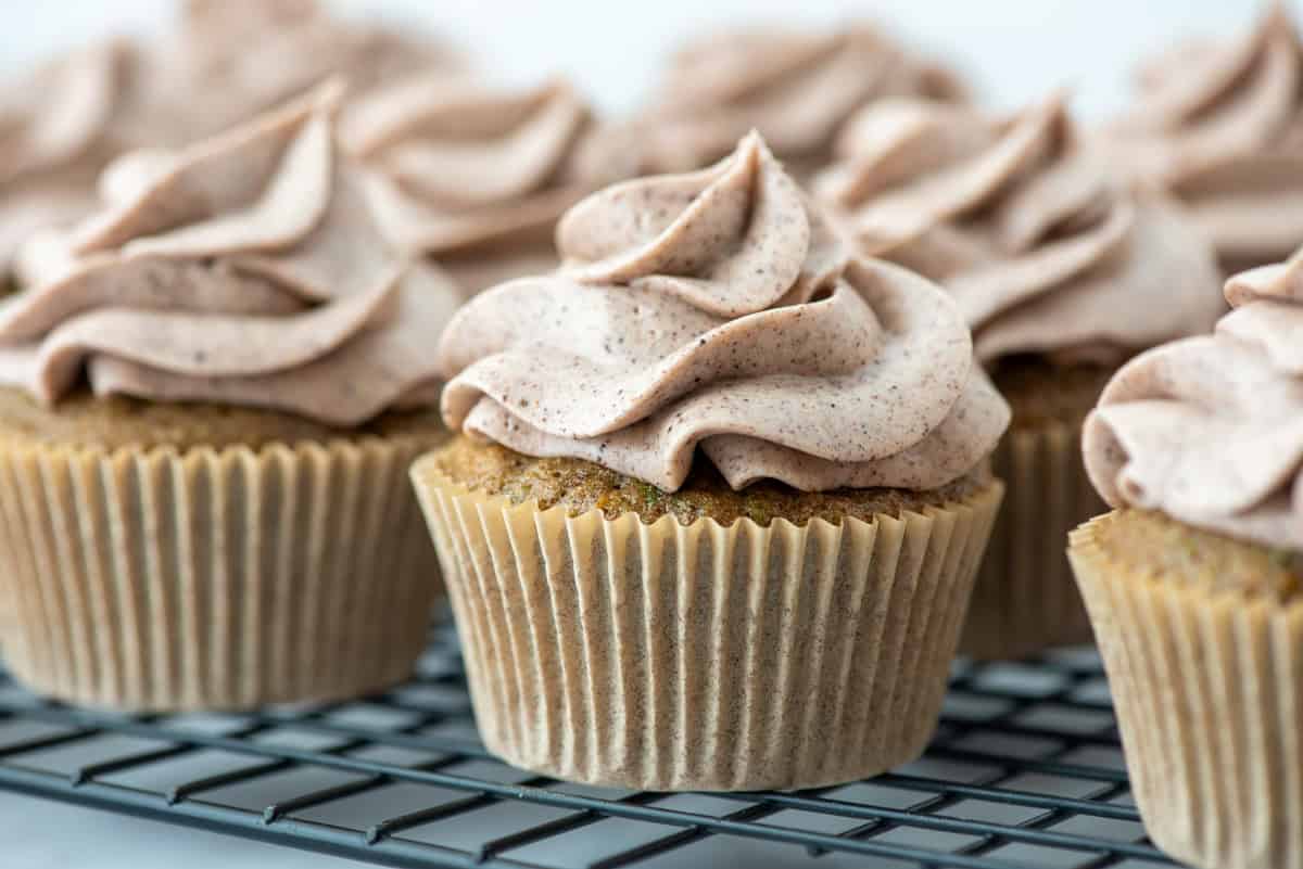 Several cupcakes with light brown frosting are arranged on a wire cooling rack, with a focus on their swirled tops and paper liners.