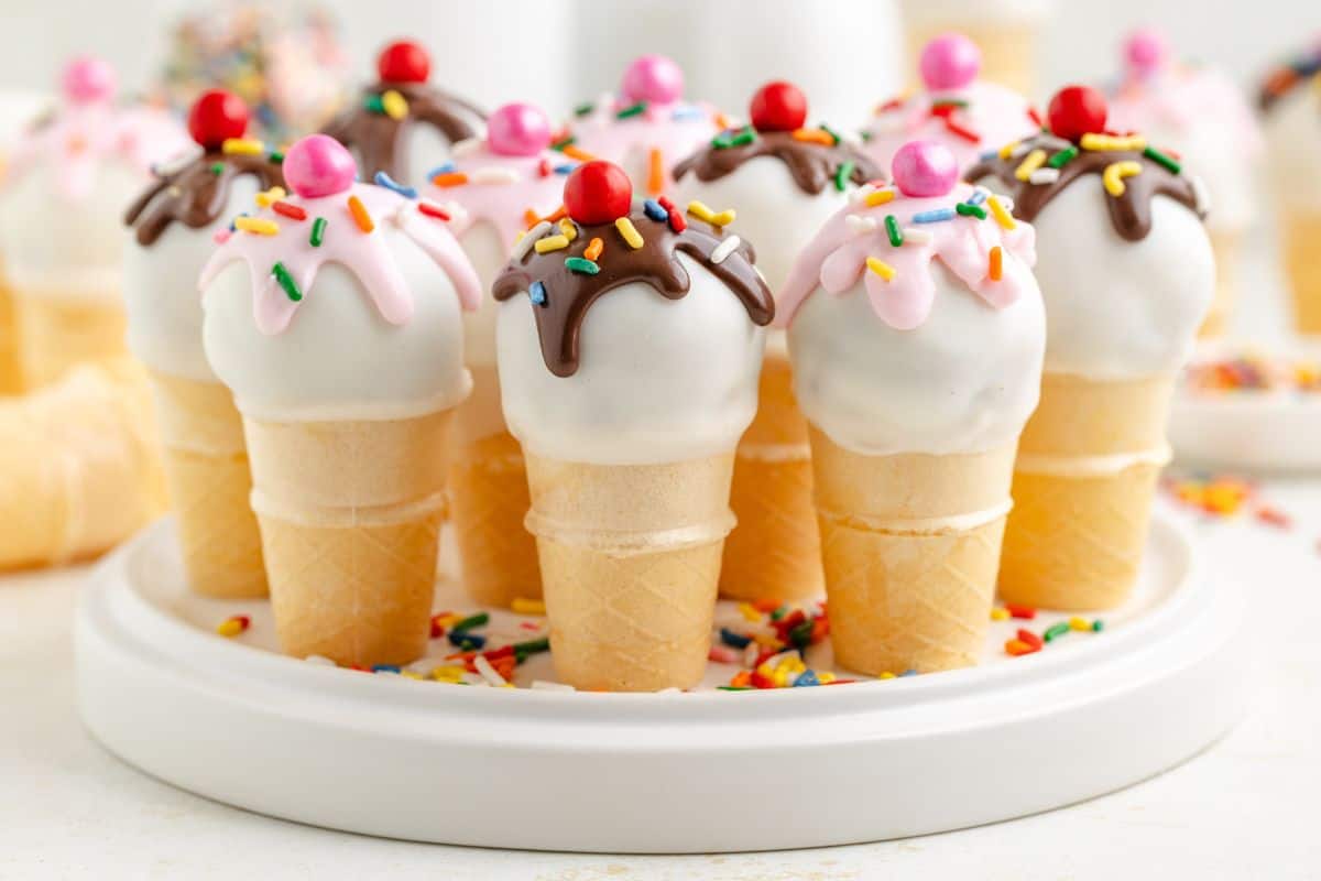 Mini ice cream cone cake pops with white, pink, and chocolate icing, topped with sprinkles and red candies, arranged on a white plate.