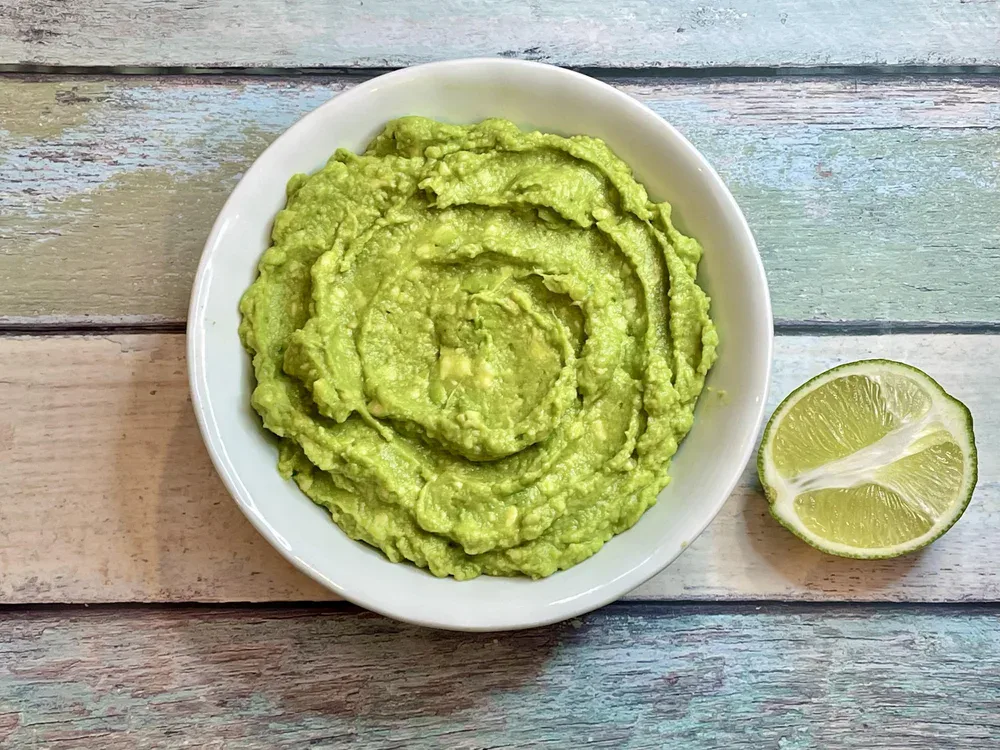 A white bowl filled with guacamole sits next to a halved lime on a rustic wooden surface.