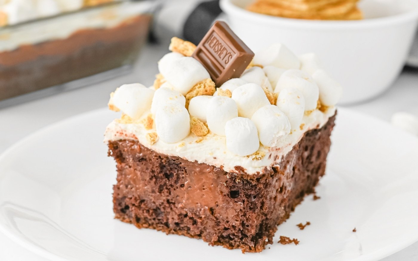 A slice of chocolate cake topped with white frosting, mini marshmallows, crumbled graham crackers, and a piece of Hershey’s chocolate on a white plate.