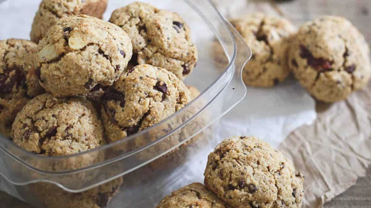 A clear container filled with several oatmeal cookies containing chocolate chips, with a few cookies placed outside on a piece of parchment paper.