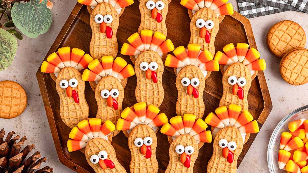A wooden plate with cookies decorated to look like turkeys using candy corn, candy eyes, and red candies; whole cookies and candy are nearby.