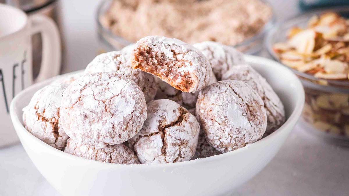 A white bowl filled with round, powdered sugar-coated cookies, with one cookie on top showing a bite taken out. Bowls of almonds and brown sugar are in the background.